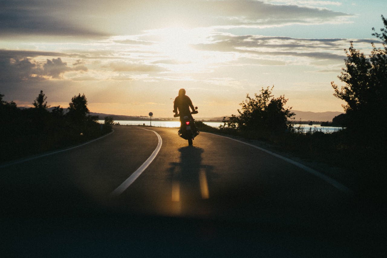 gallery-02 Silhouette of a motorcyclist riding on a scenic road during a beautiful sunset, emphasizing freedom and adventure.